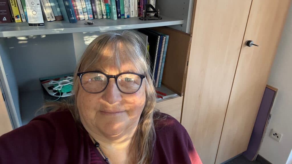 Andrea Sam vor einem Bücherregal, nachdenklich und klar, mit Lichtreflexion in der Brille – passend zum Artikel über Kommunikationstrends 2026.