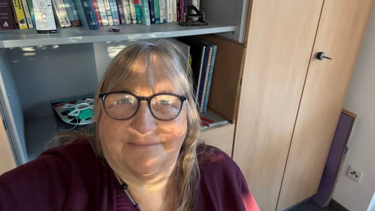 Andrea Sam vor einem Bücherregal, nachdenklich und klar, mit Lichtreflexion in der Brille – passend zum Artikel über Kommunikationstrends 2026.